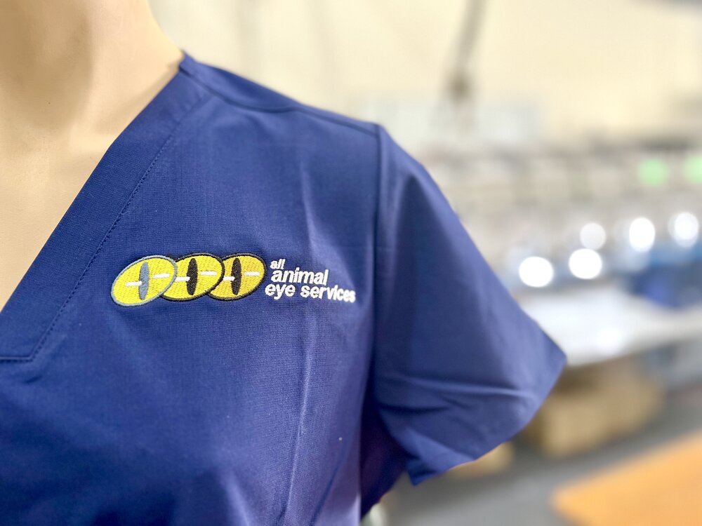 An embroidered All Animal Eye Services logo on the left chest of a navy scrub top.