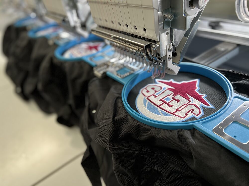 A multi-head embroidery machine part-way through stitching a club crest onto black kit bags.